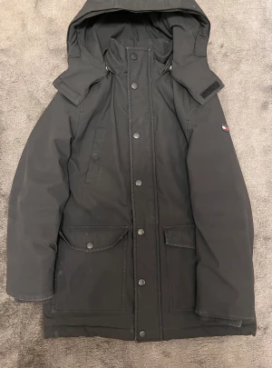 TOMMY HILFIGER JACKA - Parkajacka från Tommy Hilfiger i svart med stor logga på ryggen och ikonisk färgblockad insida i rött, vitt och blått. Jackan har huva, stora fickor med lock och knappar framtill samt dragkedja. Perfekt för kalla dagar med snygg streetstil. ( Stl: 140 Barn storlek )