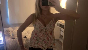 Blommig vit linnetopp med spetsdetaljer - Säljer en söt vit linnetopp med blommigt mönster i rosa, grönt och blått. Toppen har tunna axelband, v-ringad hals och spetsdetaljer med broderade hålmönster. Perfekt för varma dagar och har en feminin, luftig känsla.