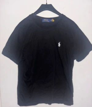 Svart t-shirt från Polo Ralph Lauren - Svart t-shirt från Polo Ralph Lauren med klassisk vit logobrodering på bröstet. Rund hals och korta ärmar, tillverkad i mjuk bomull för skön känsla. Perfekt för dig som gillar stilrena och enkla plagg med ikonisk touch. Storlek S