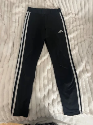 Svarta Adidas träningsbyxor  - Svarta träningsbyxor från Adidas med vita ränder längs sidorna och logga på framsidan. Byxorna är tillverkade i Primegreen-material och har en elastisk midja för skön passform. Perfekta för sport eller chill.