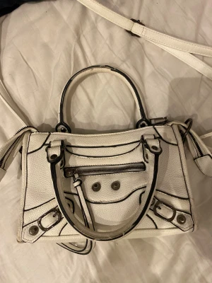 Vit handväska i skinn med svarta detaljer - Cool vit handväska i skinn med svarta kontrastsömmar och silverfärgade metalldetaljer. Väskan har både handtag och axelrem, dragkedja upptill och en mindre ficka med dragkedja framtill. Insidan är fodrad i beige tyg och har en innerficka med dragkedja.