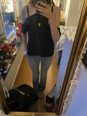 Mörkblå omlott blus med volang💕 - Säljer en mörkblå blus med omlott-design och volangdetaljer längs axlarna. Toppen har offshoulder-stil med smala axelband och en snygg v-ringning framtill. Materialet är mjukt och har ett lätt glansigt utseende, perfekt för dig som gillar trendiga detaljer!