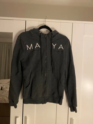 Mörkgrå hoodie från Delorez - Mörkgrå hoodie från Delorez med vit broderad text fram och bak. Tröjan har dragkedja, huva med snörning och två stora fickor framtill. Skön passform. Säljes då den är på gränsen till för liten 😊