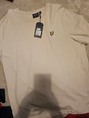 Beige t-shirt från Lyle & Scott XXL - Snygg beige t-shirt från Lyle & Scott i storlek XXL. Klassisk rund hals och korta ärmar. På bröstet finns den ikoniska broderade gula örnen. Tillverkad i mjuk bomull som känns skön mot huden. Perfekt för dig som gillar stilrena och enkla plagg.