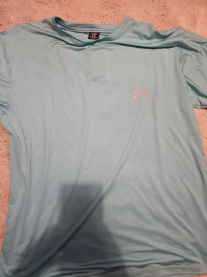 Ljusblå Under Armour t-shirt XXL - Snygg ljusblå t-shirt från Under Armour i storlek XXL. T-shirten har korta ärmar, rund hals och är tillverkad i ett lätt, ventilerande polyestermaterial. Perfekt för träning med den klassiska Under Armour-loggan tryckt på bröstet.
