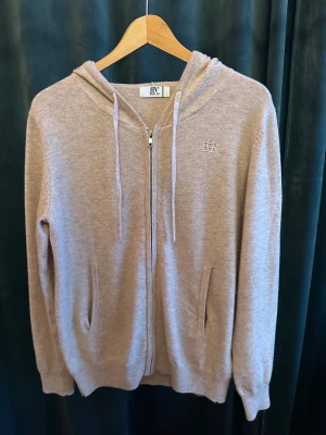 Beige hoodie med dragkedja från BVALDI - Mysig beige hoodie från BVALDI med broderad logga på bröstet. Tröjan har huva med snörning, hel dragkedja framtill och två fickor. Tillverkad i stickat material och har långa ärmar. Perfekt för lager-på-lager under höst och vinter. Helt ny, aldrig använd, nypris 1199kr, vårt pris 749kr. Str xs.