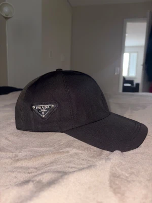 Svart Prada keps  - Snygg svart Prada keps med klassisk böjd skärm och ikonisk metallogo på sidan. Kepsen har stilren design och är tillverkad i ett slätt syntetmaterial som ger en exklusiv känsla. Perfekt för dig som vill ha en clean och lyxig look.