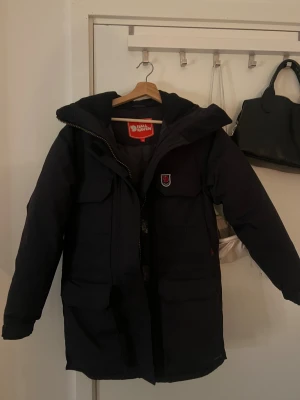 Svart Fjällräven jacka  -  Fjällräven i svart med stor huva, flera praktiska fickor och dragkedja framtill. Jackan har Fjällräven-logga på bröstet och är tillverkad i slitstarkt syntetmaterial. Perfekt för kalla dagar och har en clean, klassisk look. Kan diskutera mer om pris