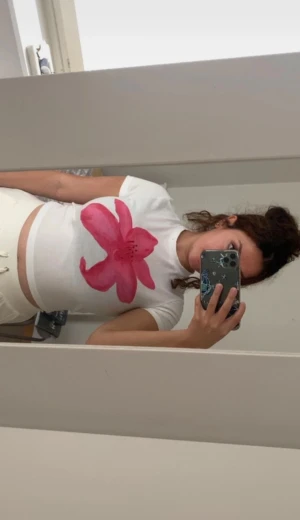 Vit t-shirt med rosa blomma - Vit kortärmad t-shirt med en stor rosa blomma tryckt framtill. Modellen är croppad och har en tight passform som framhäver midjan. Perfekt för dig som vill ha en söt och trendig look med en färgklick. Använt fåtal gånger men är fortfarande i god skick.