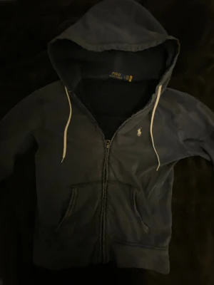 Mörkblå hoodie från Polo Ralph Lauren - Mörkblå hoodie från Polo Ralph Lauren med dragkedja, vit broderad logga på bröstet och vita snören i huvan. Klassisk känguruficka framtill och mjukt bomullsmaterial. Perfekt för chill dagar och snygg streetstil.