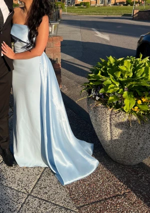 Ljusblå axelbandslös balklänning - Säljer en elegant ljusblå långklänning i glansigt satinliknande material. Klänningen är axelbandslös med en snygg omlott-detalj upptill och har en lång, svepande kjol som ger en riktigt lyxig känsla. Perfekt för bal eller speciella tillfällen! Säljer den billigare då man kan ha problem med dragkedjan.