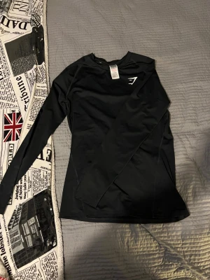 Compression Gymshark - Aldrig använd Svart långärmad träningströja från Gymshark med stilren logga på bröstet. Tröjan har en slim passform och är gjord i ett mjukt syntetmaterial som andas bra. Perfekt för gymmet eller löprundan när du vill ha något bekvämt och snyggt.
