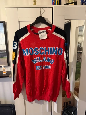 Röd Moschino tröja med tryck - Säljer en röd långärmad tröja från Moschino med stora blå och vita tryck på bröstet och coola detaljer på ärmarna. Tröjan har svarta och vita inslag på axlar och ärmar samt broderade märken och siffror. Perfekt statement piece för dig som gillar streetstyle.