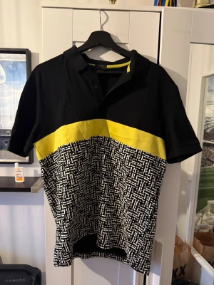 Svart och gul mönstrad pikéskjorta - Cool pikéskjorta från Sonneti i svart med en bred gul panel över bröstet och ett grafiskt vitt och svart mönster nedtill. Skjortan har korta ärmar, klassisk krage och knappar vid halsen. Perfekt för dig som gillar att sticka ut med färg och mönster.