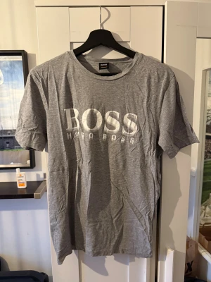 Grå t-shirt från Hugo Boss - Snygg grå t-shirt från Hugo Boss med stort vitt logotryck på bröstet. Klassisk rund hals och korta ärmar. Tillverkad i mjuk bomull som känns skön mot huden. Perfekt för dig som gillar stilrena och enkla plagg med tydlig branding.