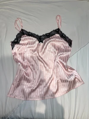 Rosa satin linnetopp med spets - Säljer en ljusrosa linnetopp i satin med tunna axelband och svarta spetsdetaljer längs halsringningen. Toppen har ett subtilt randigt mönster och en liten rosett framtill. Perfekt för dig som gillar en mjuk och glansig look.