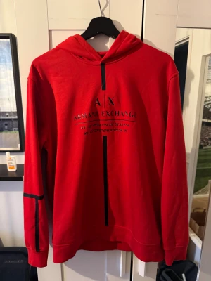 Röd hoodie från Armani Exchange - Snygg röd hoodie från Armani Exchange med svart tryck och coola svarta detaljer på ärmar och rygg. Tröjan har huva och är långärmad, perfekt för dig som vill sticka ut med en streetig look. Mjuk och skön i bomullsmix.