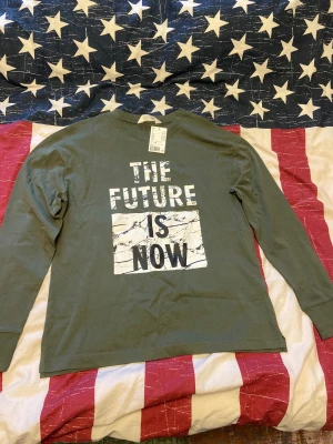 Grön långärmad tröja H&M "The Future Is Now" - Cool grön långärmad tröja från H&M med trycket "THE FUTURE IS NOW" i vitt och svart, samt marmormönster i bakgrunden. Perfekt för dig som gillar statement-plagg och streetstyle. Skön passform och tillverkad i mjuk bomull. Tunn tröja, nästan som en långärmad T-shirt. Storlek 170 barn. Prislappen är fortfarande på, alltså den är helt ny och inte använd. 