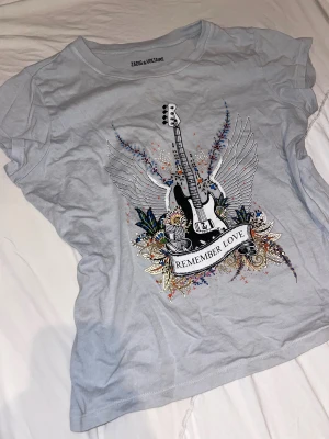 Ljusblå t-shirt Zadig & Voltaire - Snygg ljusblå t-shirt från Zadig & Voltaire med coolt tryck av en elgitarr, vingar och blommor samt texten 'Remember Love' på framsidan. T-shirten har korta ärmar och är gjord i mjuk bomull. Perfekt för dig som gillar Zadig & Voltaire.