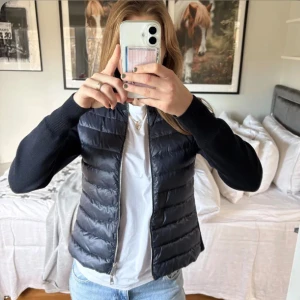 Svart Moncler pufferjacka med stickade ärmar - Säljer en svart pufferjacka från Moncler med stickade ärmar och quiltad framsida. Jackan har dragkedja och Moncler-logga på ärmen. Perfekt för dig som vill ha en snygg och chill look med exklusiv känsla. Passar till jeans och sneakers.