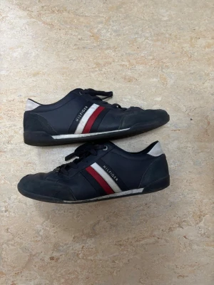 Tommy Hilfiger Corporate - Snygga mörkblå sneakers från Tommy Hilfiger med klassiska röda och vita ränder på sidan och Hilfiger-logga. Skorna har rund tå, platt sula och snörning. Materialet är skinn med detaljer i syntet och mocka. Perfekt för dig som gillar stilrena och sportiga sneakers.