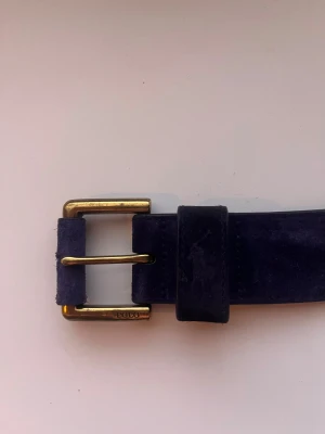 Mörkblå mockabälte från Polo Ralph Lauren - Snyggt mörkblått bälte i mocka från Polo Ralph Lauren med guldfärgat spänne där det står 'POLO' ingraverat. Bältet är brett och har en klassisk, stilren design som passar till jeans eller kostymbyxor.