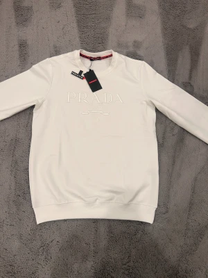 Vit sweatshirt från Prada - Exklusiv vit sweatshirt från Prada med broderad logga och Milano-text framtill. Tröjan har rund hals, långa ärmar och ribbade muddar. Snygg detalj med röd logoband på insidan av kragen. Perfekt för dig som gillar stilrena designer och high fashion. Storlek m men passar s också.