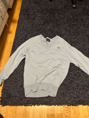 Ljusblå v-ringad tröja Fred Perry - Snygg ljusblå v-ringad tröja från Fred Perry med broderad logga på bröstet. Tröjan är stickad i mjukt material och har långa ärmar samt ribbade muddar vid ärmslut och nederkant. Perfekt för dig som gillar klassisk stil med en modern touch.