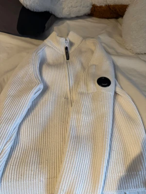 Vit ribbad kofta från C.P. Company -  Ny pris 2,7 det va rea Säljer en vit ribbstickad kofta från C.P. Company perfekt i vintern med dragkedja framtill och deras ikoniska linsdetalj på ärmen. Koftan har hög krage och är långärmad, perfekt för lager-på-lager. Materialet är mjukt och stickat, vilket ger en clean och modern look. Har ett lite defekt skriv så skickar jag