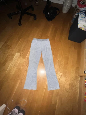 Grå mjukisbyxor juicy couture - Säljer ett par grå mjukisbyxor med bootcut-ben och snörning i midjan. Byxorna har fickor både fram och bak samt ett broderat motiv på ena sidan. Materialet är mjukt och skönt, perfekt för chill dagar. Passformen är loose och midjan är hög. Det är också äkta  juciy couture som är dyrt men jag säljer det för lågt pris, det är i bra skick men bilderna blev inte så bra.