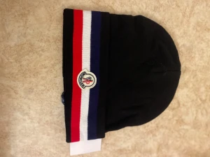 Moncler mössa med ränder - Snygg svart mössa från Moncler med breda randiga detaljer i rött, vitt och marinblått. Framtill sitter Monclers klassiska broderade logotyp. Mössan är stickad och har en mjuk, skön känsla som passar perfekt när det är kallt ute.
