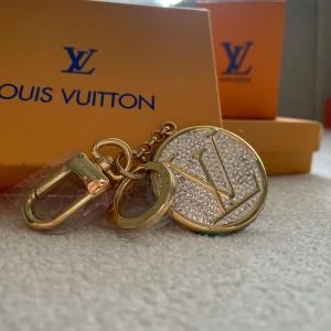  Vuitton glittrig nyckelring - Exklusiv rund nyckelring från  Vuitton i guldtonad metall med -logga framtill. Framsidan är täckt av glittrande stenar som ger en lyxig vibe. Baksidan är blank med ingraverad  Vuitton Paris. Perfekt accessoar för att lyfta din väska eller nycklar.