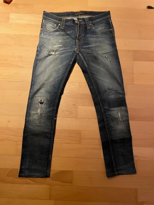 Blå slitna jeans från Nudie Jeans - Säljer ett par blå jeans från Nudie Jeans med snygga slitningar och lagade detaljer på benen. Klassisk femficksmodell med orange sömmar och Nudie-logga på bakfickan. Jeansen har rak passform och är tillverkade i jeansmaterial med dragkedja.