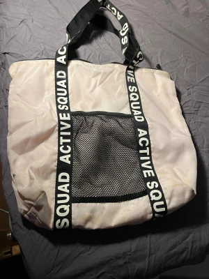 Vit shoppingväska ACTIVE SQUAD - Stor vit shoppingväska från ACTIVE SQUAD med svarta handtag och text i vitt. Väskan har ett stort nätfack framtill och dragkedja upptill. Perfekt för gymmet eller när du behöver extra plats till dina grejer.