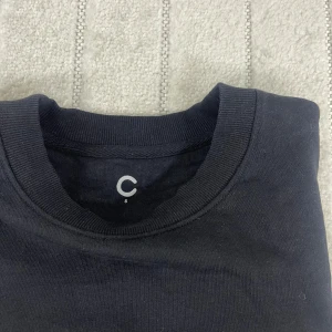 Svart crewneck från C, storlek S - En stilren svart crewneck-tröja från C i storlek S. Tröjan har rund halsringning, långa ärmar och ribbade muddar vid ärmslut och nederkant. Perfekt basic-plagg i mjuk bomull som funkar året runt.