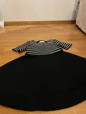 Randig paljettklänning Holly & Whyte - Svart och vit randig klänning från Holly & Whyte med paljettdetaljer upptill. Klänningen har trekvartsärm och en svart, utsvängd kjol. Perfekt för dig som gillar lite extra bling och klassisk stil.