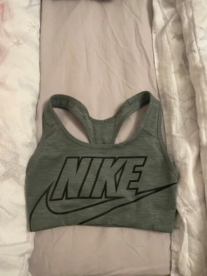 Grön sporttopp från Nike Dri-Fit XS - Säljer en grön sporttopp från Nike i storlek XS med stort svart Nike-tryck framtill. Modellen är ärmlös med brottarrygg och tillverkad i Dri-Fit polyester som andas. Perfekt för träning och har en tight passform som sitter snyggt på kroppen.