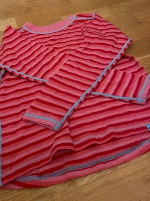 Randig långärmad tröja Pierre Robert - Säljer en långärmad tröja från Pierre Robert i rosa och rött med randigt mönster. Tröjan har ljusblå sömmar som snygg detalj och är tillverkad i mjukt material, perfekt för lager-på-lager under höst och vinter.