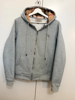 Burberry Hoodie - Grå hoodie från Burberry med klassiskt rutigt foder i huvan och broderad logga på bröstet. Tröjan har dragkedja framtill, snörning vid huvan och två fickor. Tillverkad i mjuk bomullsmix, perfekt för chill dagar. Snyggt ribbade muddar och normal passform.