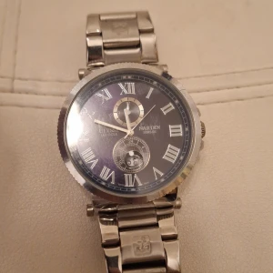 Ulysse Nardin silverfärgad herrklocka - Snygg herrklocka från Ulysse Nardin med rund boett och länkarmband i silverfärgat metall. Svart urtavla med romerska siffror, datumvisning och extra subdial. Ankare-detalj på armbandet ger en nautisk vibe. Perfekt för dig som gillar klassisk och stilren design. Batteri behövs byts