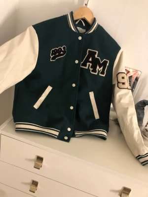 Grön och vit bomberjacka med broderi - Snygg collegeinspirerad bomberjacka från H&M i mörkgrönt med vita ärmar och detaljer. Jackan har broderade patches med 'AM' och '93' samt randiga muddar och knappar framtill. Perfekt för dig som gillar streetstyle och sportig vibe.