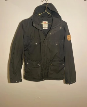 Fjällräven Greenland women S - Svart vindjacka från Fjällräven, modell Greenland W. Jackan har huva, flera fickor med tryckknappar och klassisk Fjällräven-logga på ärmen. Tillverkad i slitstark bomull och polyesterblandning, perfekt för dig som gillar outdoor och streetwear.