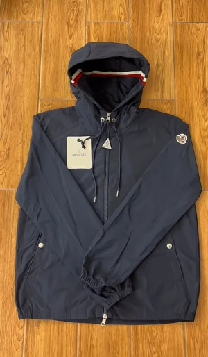 Mörkblå vindjacka från Moncler - Snygg mörkblå vindjacka från Moncler med huva och snörning. Jackan har dragkedja framtill, silverfärgade knappar på fickorna och Moncler-logga på ärmen. Insidan av huvan har en röd, vit och blå rand. Perfekt för dig som gillar stilrena och sportiga plagg.