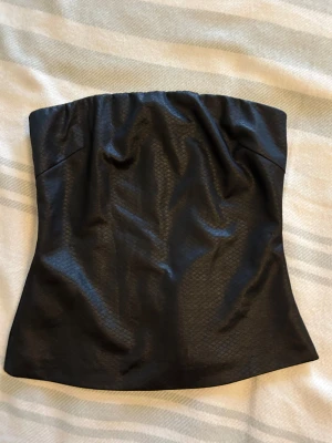 Ny Svart tubtopp i skinnlook från MNG Collection - Snygg svart tubtopp från MNG Collection i storlek S. Toppen har en cool skinnlook med diskret mönster och dragkedja bak. Den är axelbandslös och har en tight passform som sitter snyggt. Perfekt för dig som vill ha en edgy vibe på outfiten.