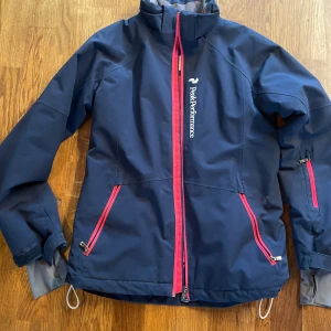 Mörkblå skidjacka Peak Performance 160 - Peak Performance skidjacka i mörkblått med snygga rosa dragkedjor och logga på bröstet. Jackan har hög krage, långa ärmar och flera praktiska fickor med dragkedja.