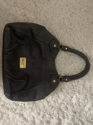 Svart axelväska Marc by Marc Jacobs - Snygg svart axelväska från Marc by Marc Jacobs i mjukt skinn med guldfärgade metalldetaljer. Väskan har en avtagbar axelrem och ett coolt innerfoder med stora bokstäver. Perfekt storlek för allt du behöver och stilren design med logoplatta framtill.