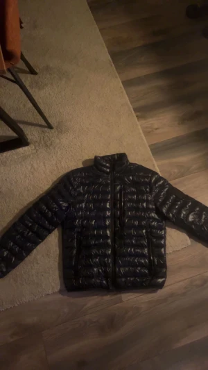 Mörkblå peak performance pufferjacka med dragkedjor - Snygg mörkblå pufferjacka med glansig yta och horisontella quiltade partier. Jackan har hög krage, tre praktiska fickor med dragkedja och långärmade ärmar. Perfekt för kalla dagar och enkel att matcha med din stil.