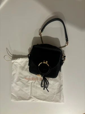 Svart axelväska från See By Chloé - Snygg svart axelväska från See By Chloé i skinn med guldfärgade metalldetaljer och dekorativ ring framtill. Väskan har kedjerem och en liten tofsdetalj. Kommer med dustbag.