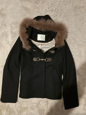 Svart kappa med päls från ef-dé - Snygg svart kappa från ef-dé med fluffig brun päls runt huvan och guldfärgade spänndetaljer framtill. Jackan har två fickor, lång ärm och är tillverkad i ullmaterial med slät, ljus insida. Perfekt för dig som gillar klassisk stil med en twist.