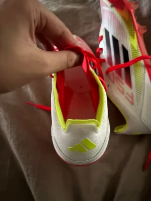 Adidas vita och neon fotbollsskor - Säljer ett par Adidas fotbollsskor med vit ovandel, neon-gula detaljer och röda snören. Skorna har inga sulor  röda och gula dobbar för gräsplan. Snyggt mönstrad mesh och klassiska svarta Adidas-ränder på sidan. Perfekt för dig som vill sticka ut på planen.